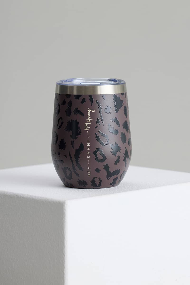 A stainless steel insulated cup with a dark brown and black leopard print pattern, featuring a clear lid and vertical branding text, displayed on a white block against a neutral gray background.