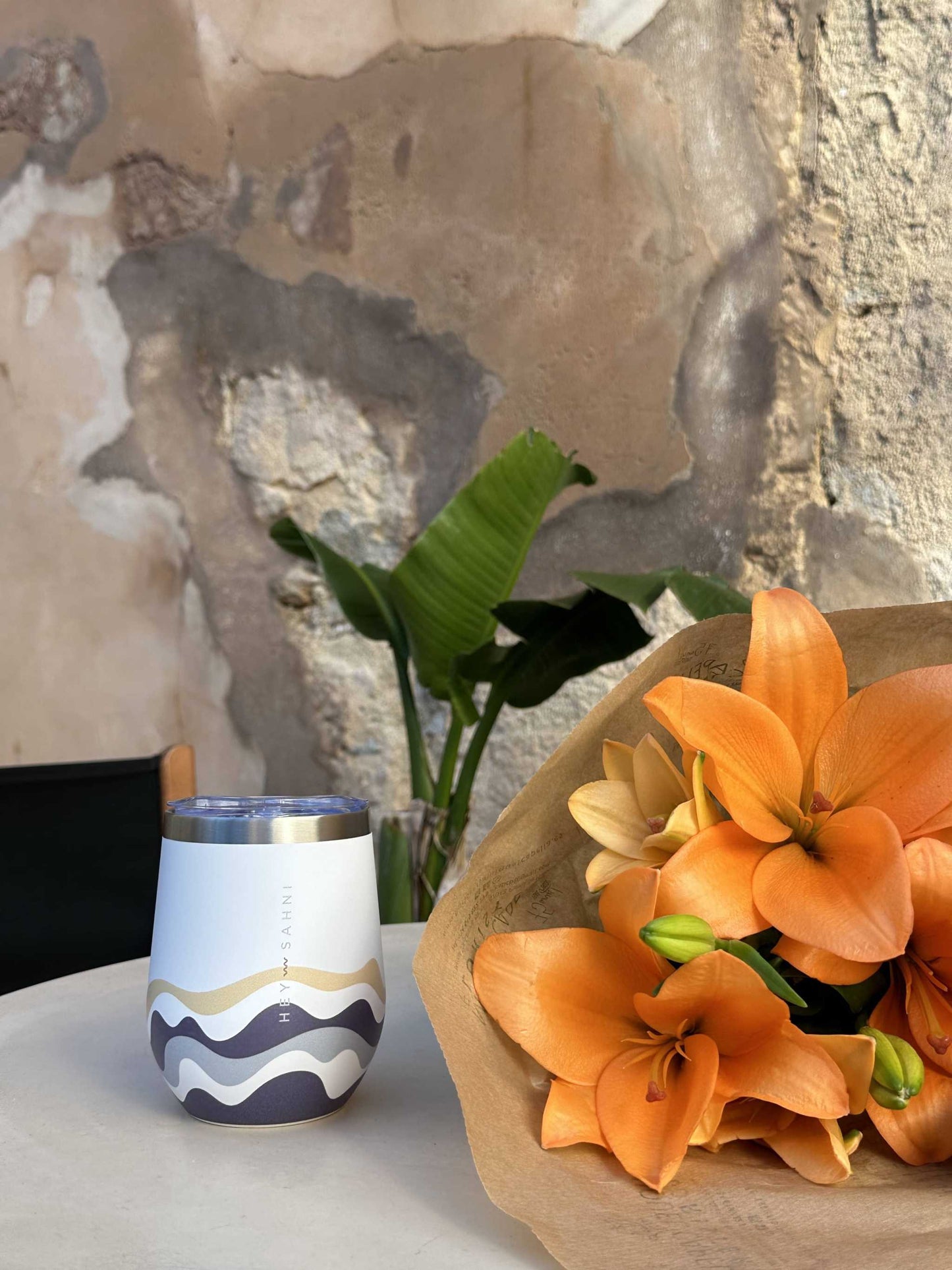 Stainless steel cup with wave design on a table, next to orange flowers, offering eco-friendly hot and cold drink storage.