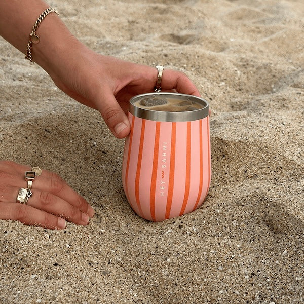 A coral pink and peach vertically striped insulated stainless steel HEY SAHNI Cup with a silver rim, held by a hand on a sandy beach.