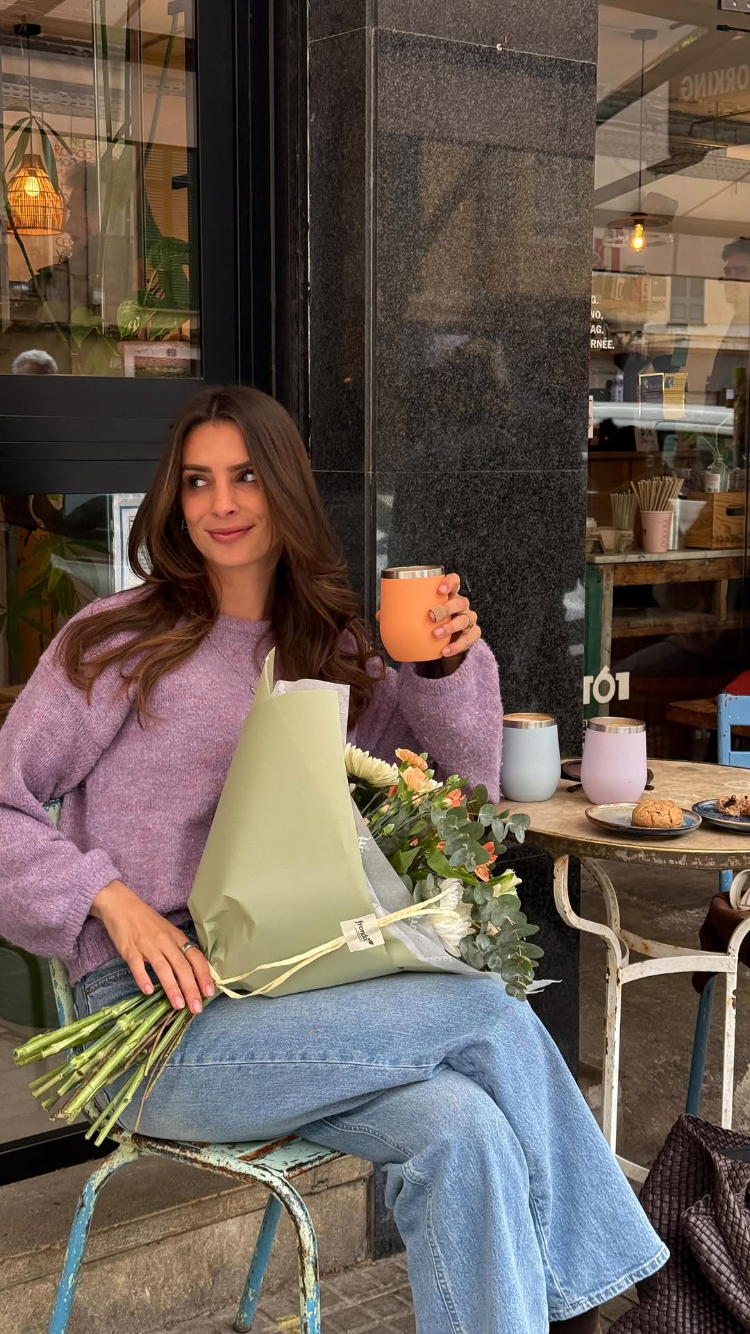 Woman holding an orange HEY SAHNI stainless steel coffee cup in a cozy café setting