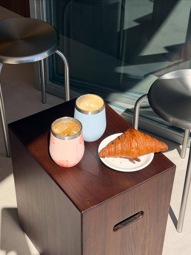 Two drinks in stainless steel coffee cups and a croissant on a wooden table with chairs in the background.