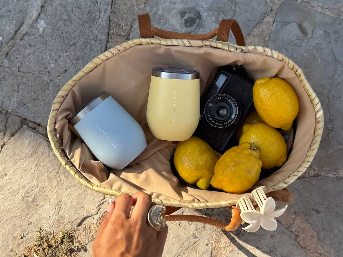 Wicker basket with lemons, a camera, and two HEY SAHNI Cups on a stone surface