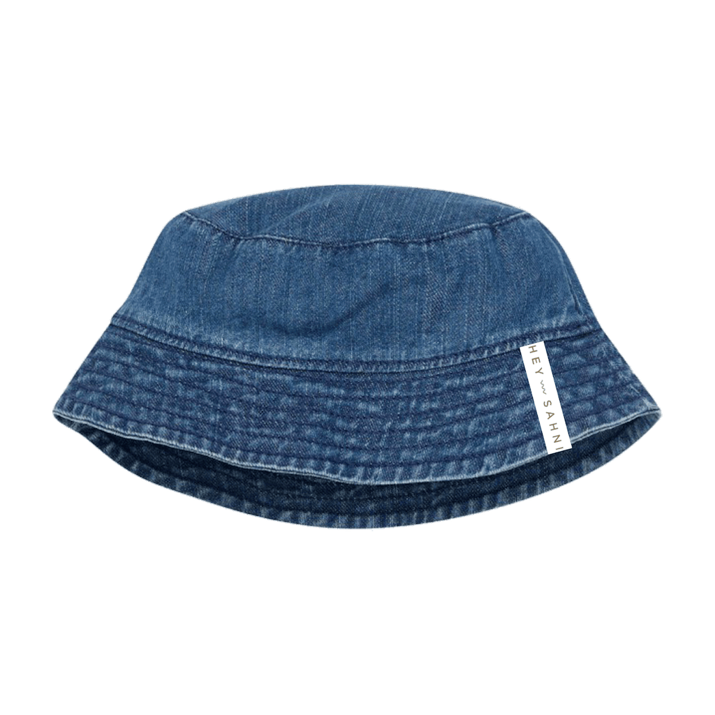 A blue denim HEY SAHNI bucket hat with a white fabric brand tag on the side, shown against a solid green background.