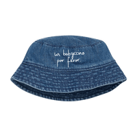 A blue HEY SAHNI denim bucket hat with white cursive text reading 'un babuccino por favor.' on the front, shown against a solid green background.