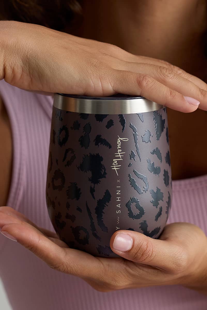 A close-up of a person holding a dark gray leopard-print insulated tumbler with a stainless steel rim, wearing a light pink ribbed top.