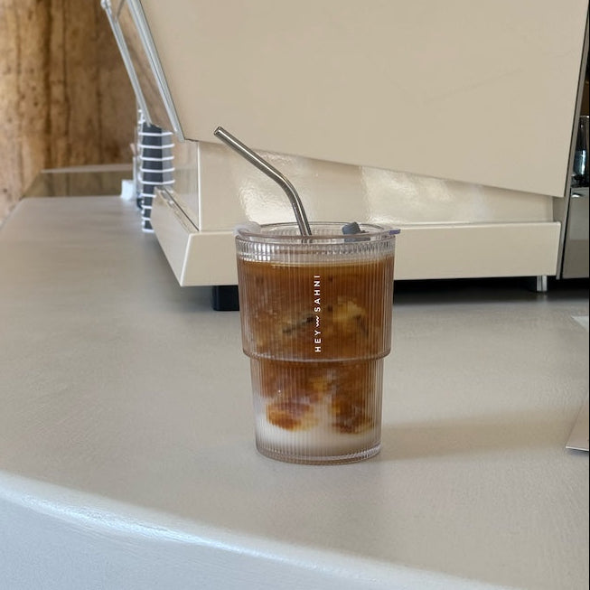 An iced coffee drink in a clear plastic cup with a metal straw sits on a white countertop in front of a cream-colored coffee machine with brown capsules visible on top.