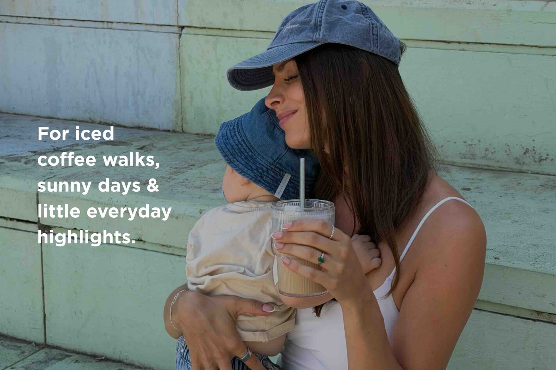 Woman wearing a blue cap holds a child in a blue bucket hat and a HEY SAHNI Iced To-Go Cup. Text reads: “For iced coffee walks, sunny days & little everyday highlights.”