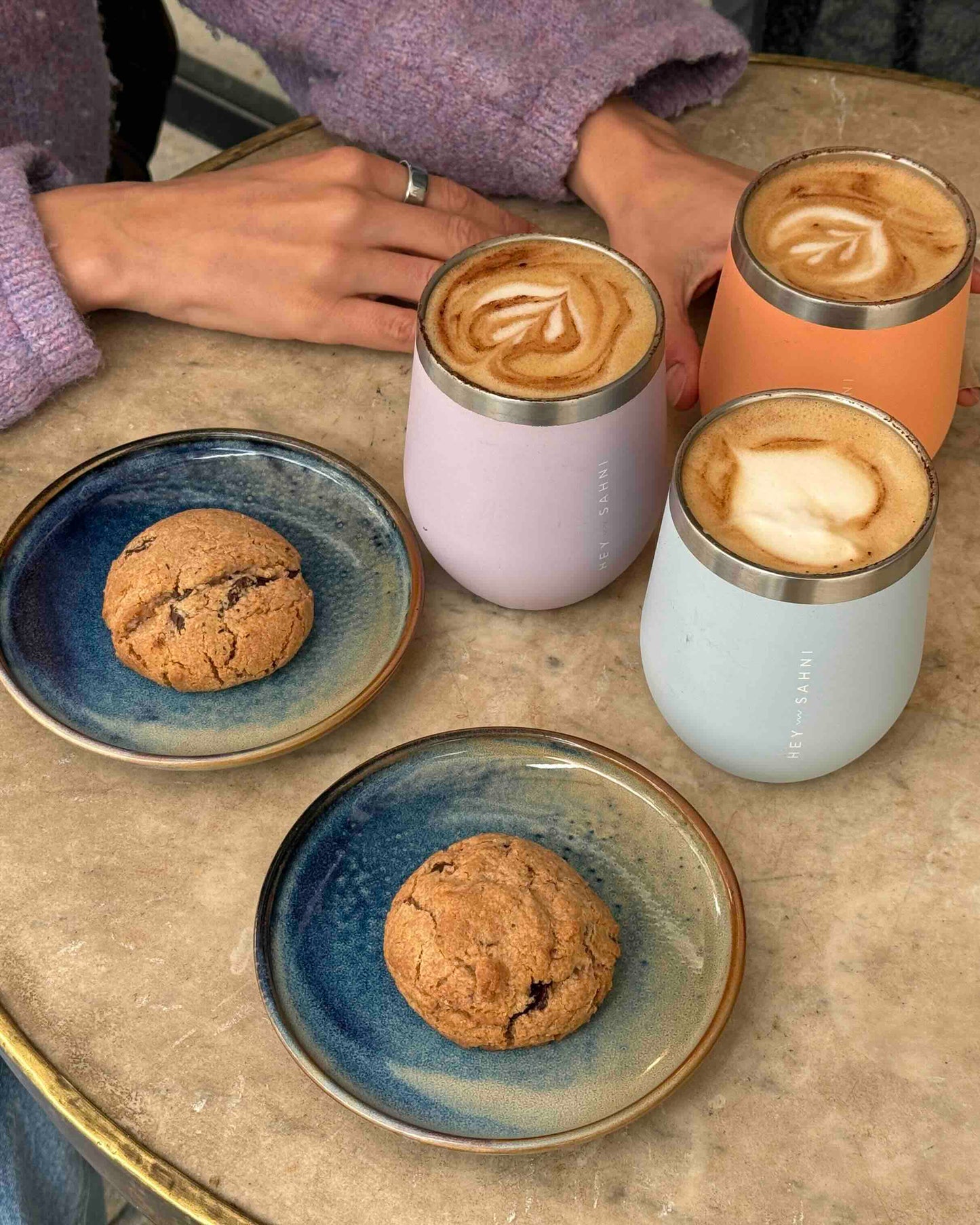 Three reusable coffee cups in soft spring colors filled with latte art, placed on a table with cookies in a cozy café setting.