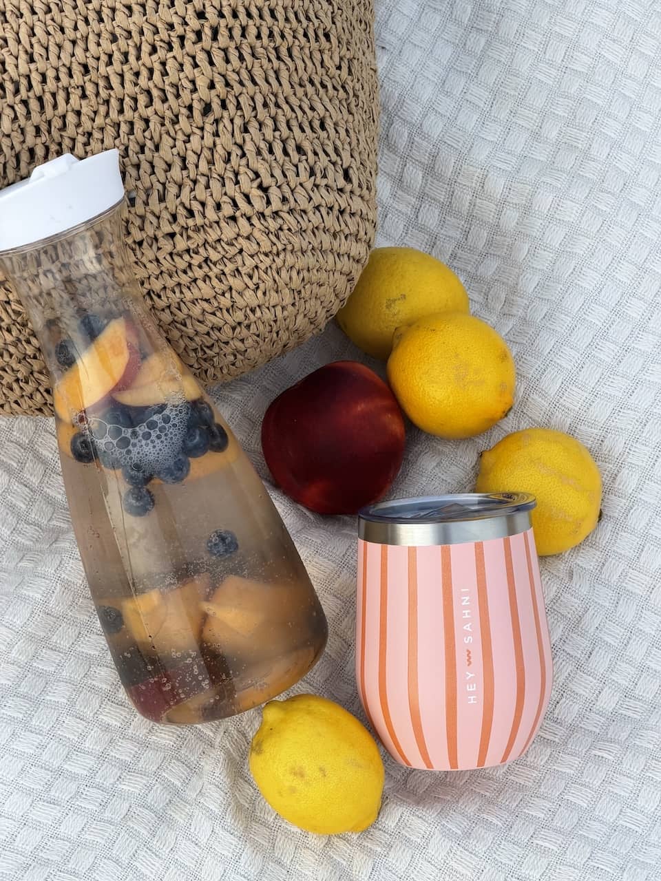 A styled flat-lay featuring a clear glass bottle filled with fruit and lemonade and a pink and white striped insulated HEY SAHNI Cup with a silver rim, arranged on a textured white blanket with lemons, a peach, and a woven basket in the background.