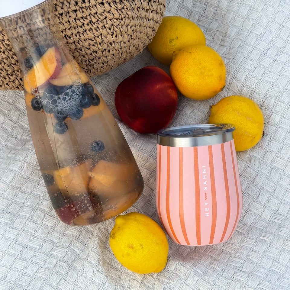 A pink and peach striped insulated Hey Sahni striped cup with a stainless steel lid sits on a textured white blanket alongside a glass bottle of iced fruit drink, lemons, a peach, and a woven basket.